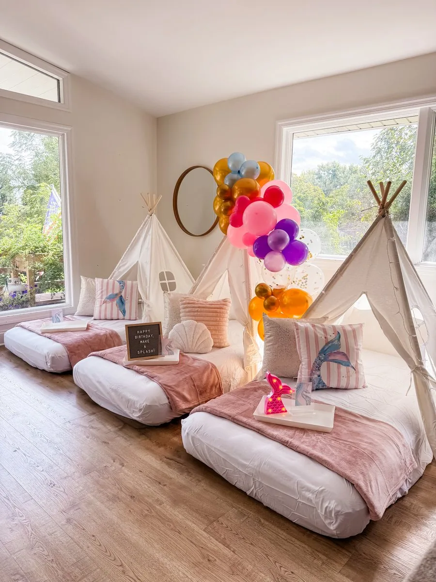 Mermaid teepee sleepover setup with white teepees, mermaid-tail pillows, blush blankets, a Happy Birthday Make a Splash sign, and a pink, purple, and gold balloon garland