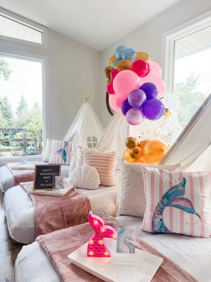 Close-up of a mermaid sleepover with a light-up mermaid tail, watercolor mermaid-tail pillow, seashell pillow, and a colorful balloon arch