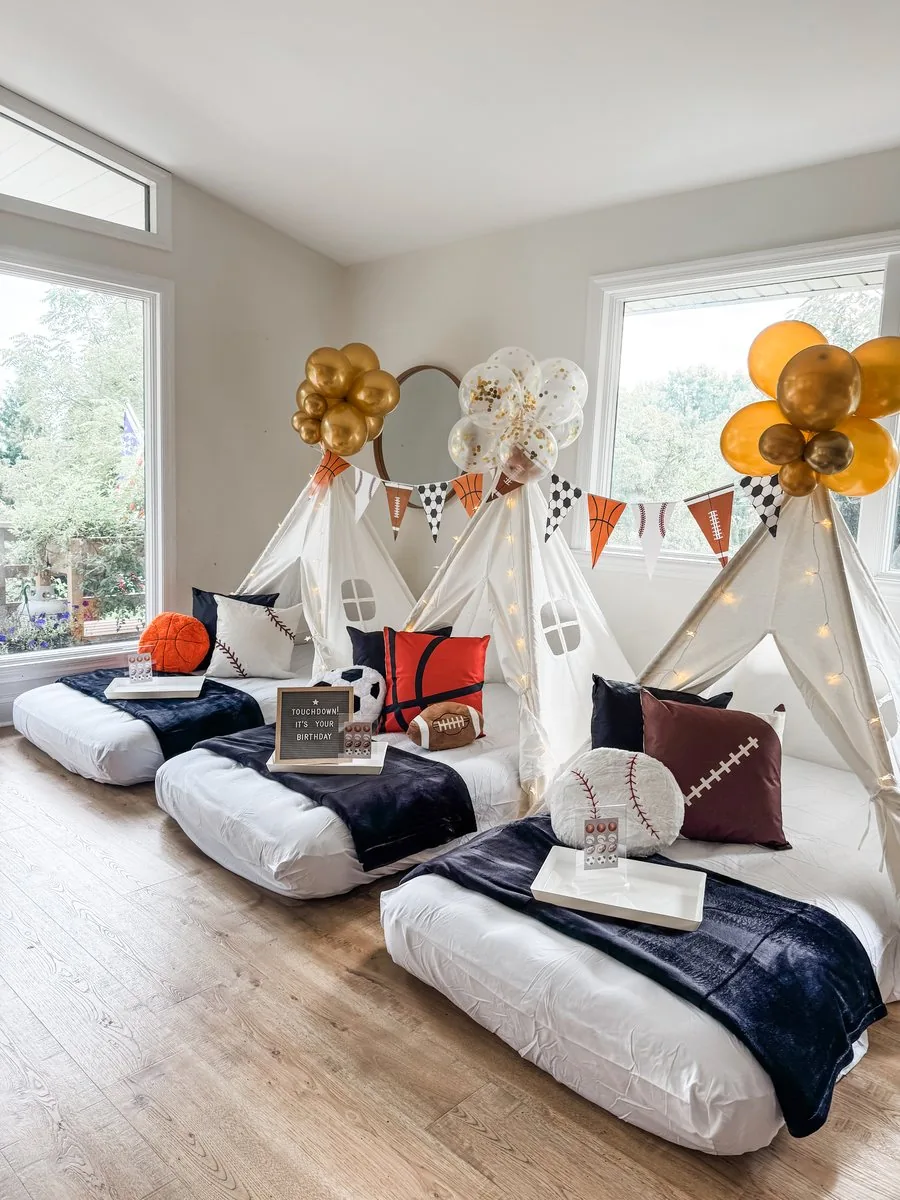 Sports-themed teepee sleepover with white teepees, navy blankets, basketball, football, and baseball pillows, a sports pennant banner, and gold balloons